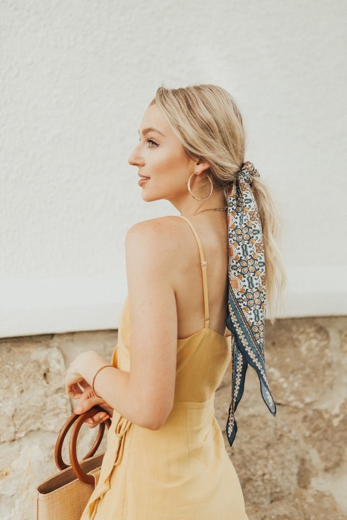 Low blonde ponytail tied with a patterned scarf, yellow slip dress and woven bag