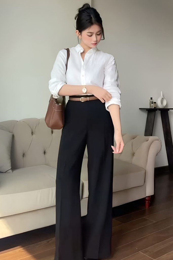 Woman in a white button up shirt with rolled sleeves, a thin belt and black high waisted wide leg trousers, holding a brown shoulder bag