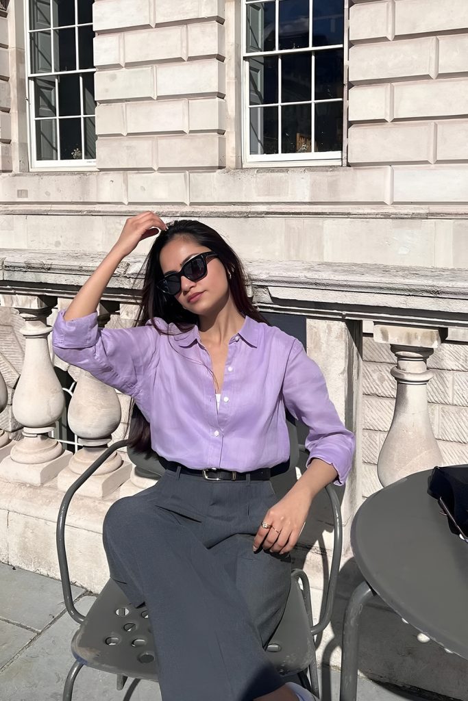 Woman in sunglasses wearing a lavender button up shirt, a black belt and grey trousers, seated beside a stone balustrade