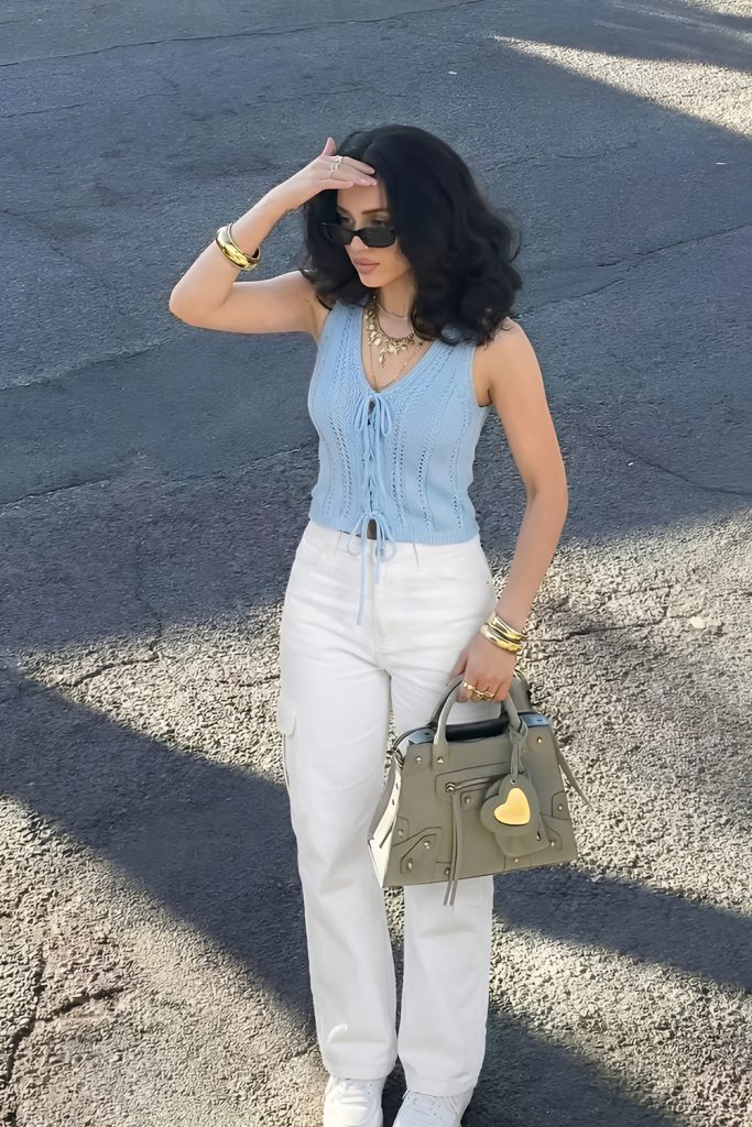 Woman in sunglasses wearing a light blue sleeveless tie front knit top and white trousers carrying a grey green handbag
