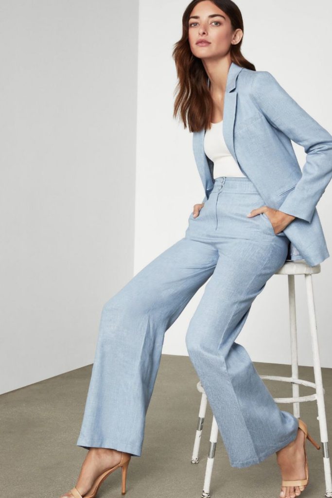 Woman seated on a white stool in a light blue blazer and matching wide-leg trousers with a white top and tan heeled sandals