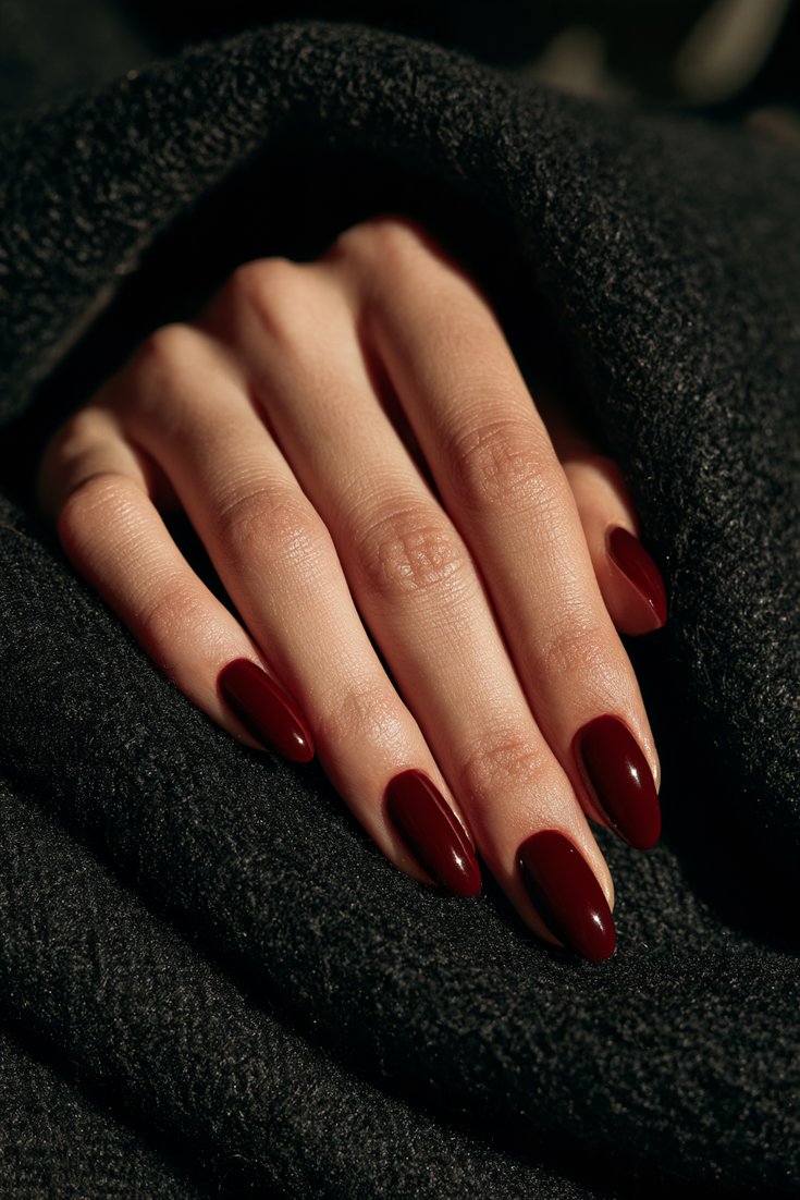 Medium almond nails in glossy deep oxblood red on a hand resting on black wool fabric