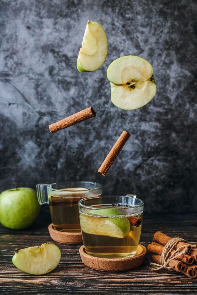 Two glasses of a spiced apple drink with apple slices and cinnamon sticks on a dark wooden table