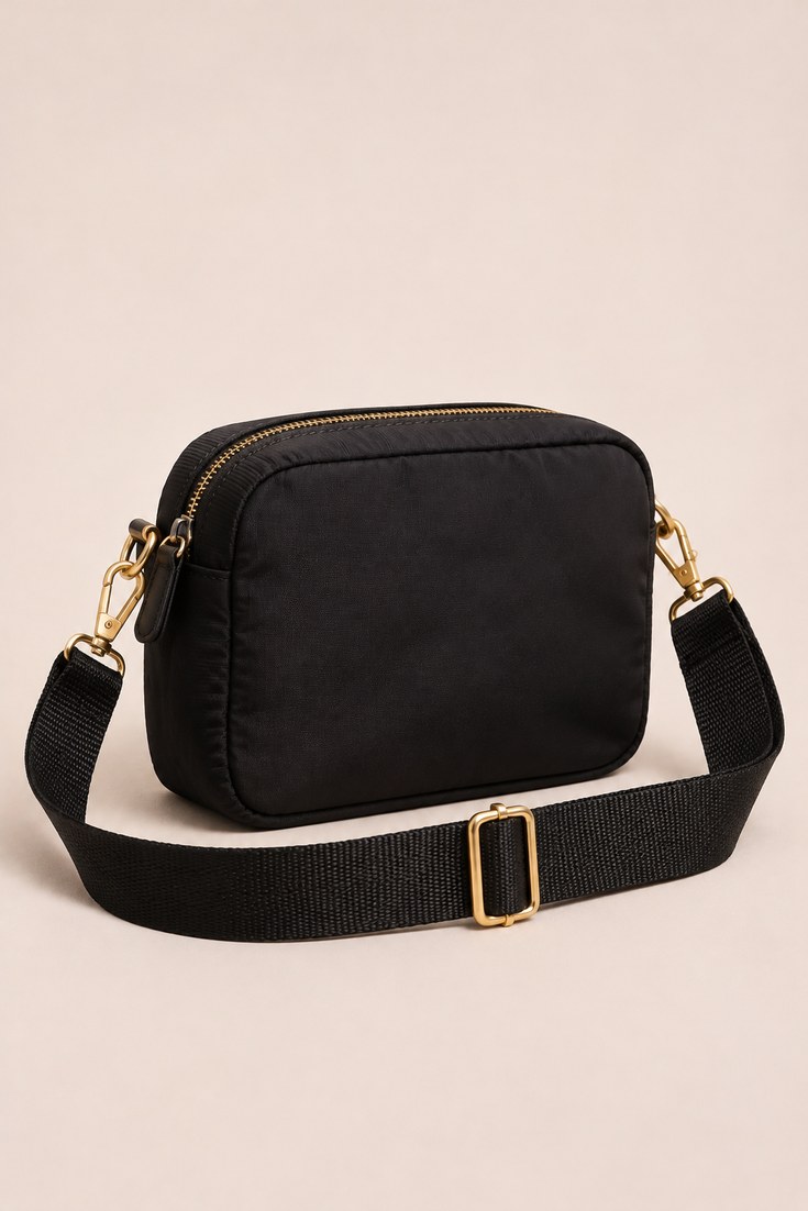 Boxy black nylon camera crossbody bag with wide webbing strap on a soft ivory backdrop