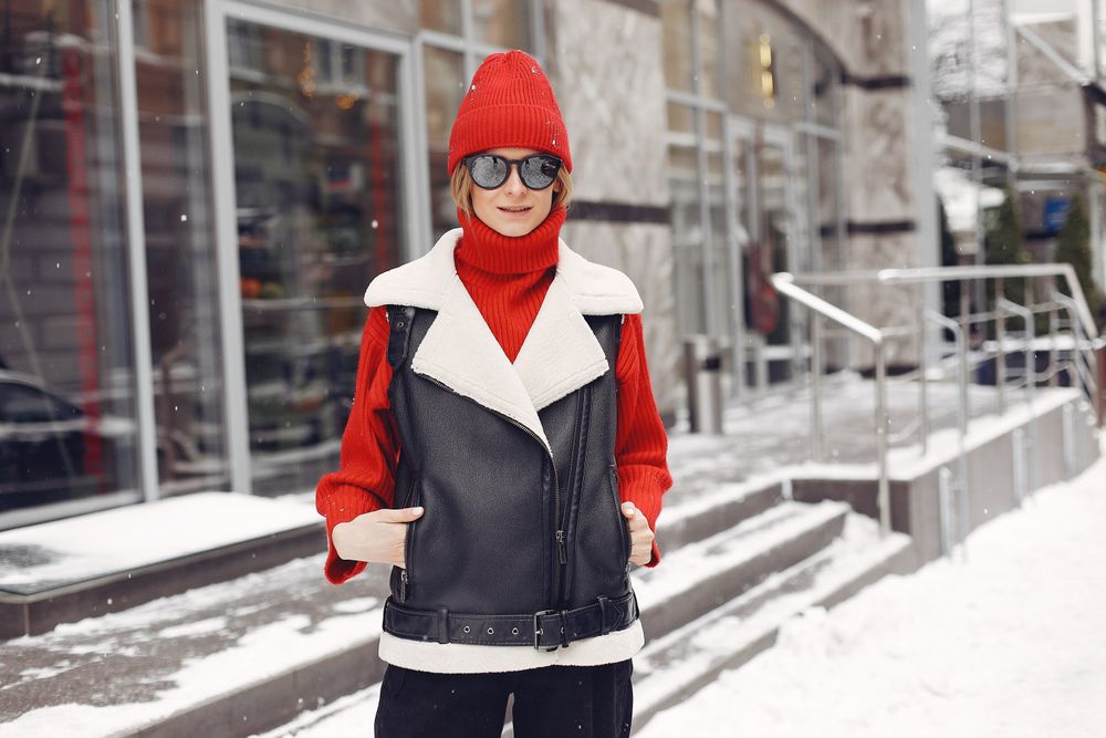 Woman in a red beanie, sunglasses, a red high-neck sweater and a black leather vest with a cream shearling collar standing on snowy steps