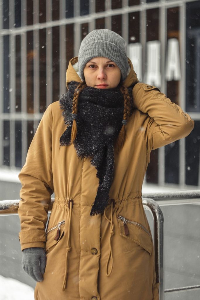 Woman in a grey beanie, a black fringed scarf and a mustard yellow parka with gloves and braids in falling snow