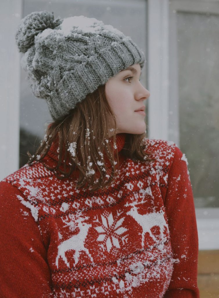 Woman in a grey cable-knit pom beanie and a red Nordic sweater with white reindeer looking to the side