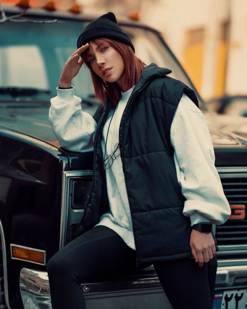 Woman in a navy quilted vest over a cream sweater leaning on a vintage car