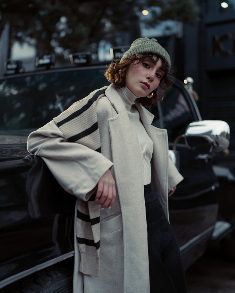 Woman in a sage green beanie and a long pale grey-green coat with a striped scarf leaning on a dark car