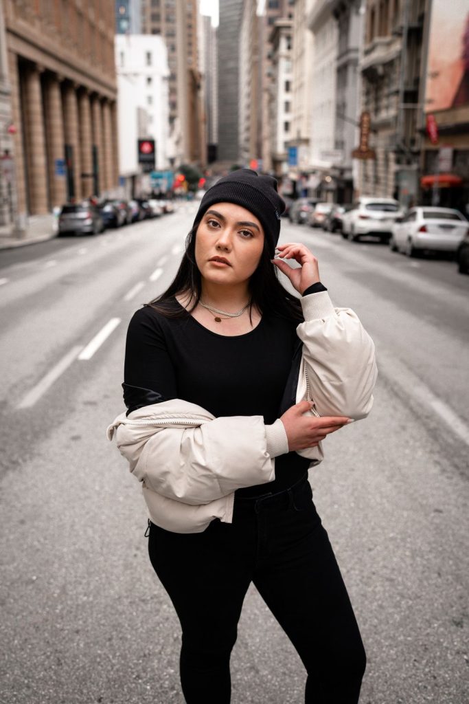 Woman in a black beanie and black top and trousers holding a cream puffer jacket over one arm on an empty city street