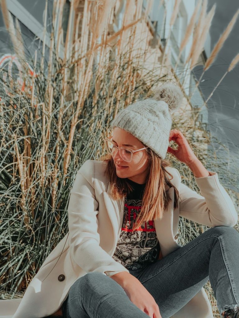 Woman in glasses, a light grey knit beanie, a cream coat, a graphic tee and jeans sitting among dry grasses