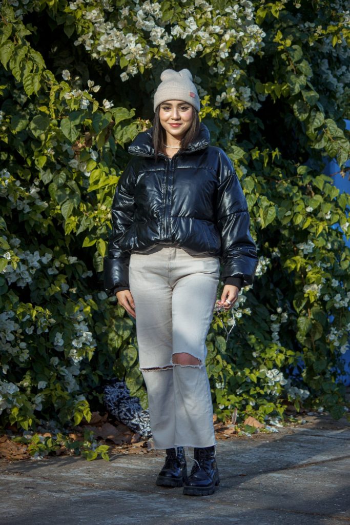 Woman in a light grey beanie, a glossy black cropped puffer jacket, light ripped jeans and black boots in front of a green hedge