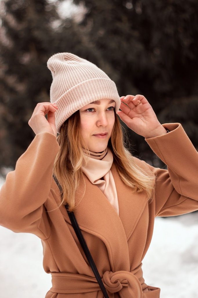 Woman in a blush pink knit beanie, a camel coat and a pale scarf standing in snow with a crossbody strap