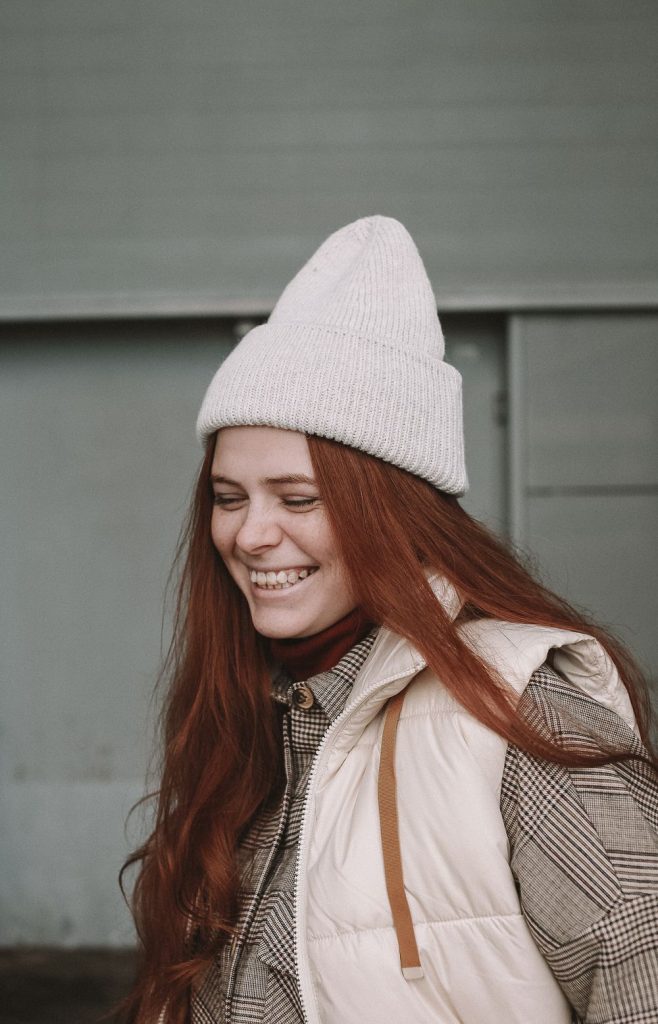 Smiling woman with long red hair in a light grey beanie and a white quilted vest over a checked coat
