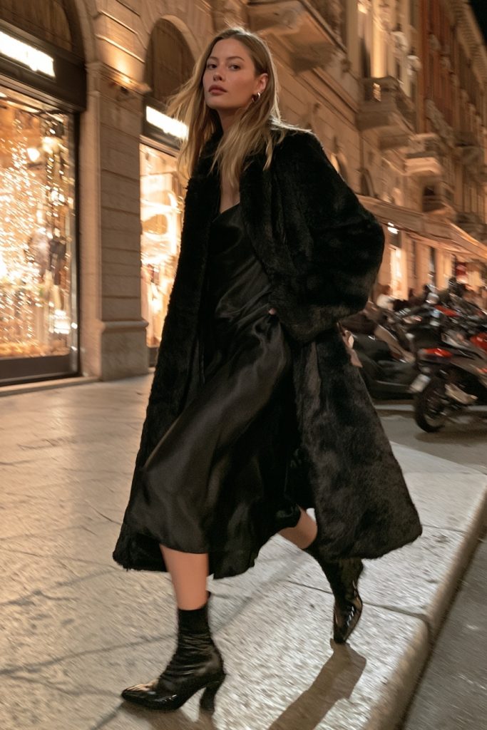 Woman in a long black faux fur coat over a black satin dress and black boots on an evening street lit by shop windows