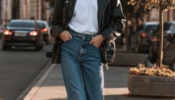 Woman in a black leather jacket over a white tee with blue jeans and white sneakers on a sunny sidewalk