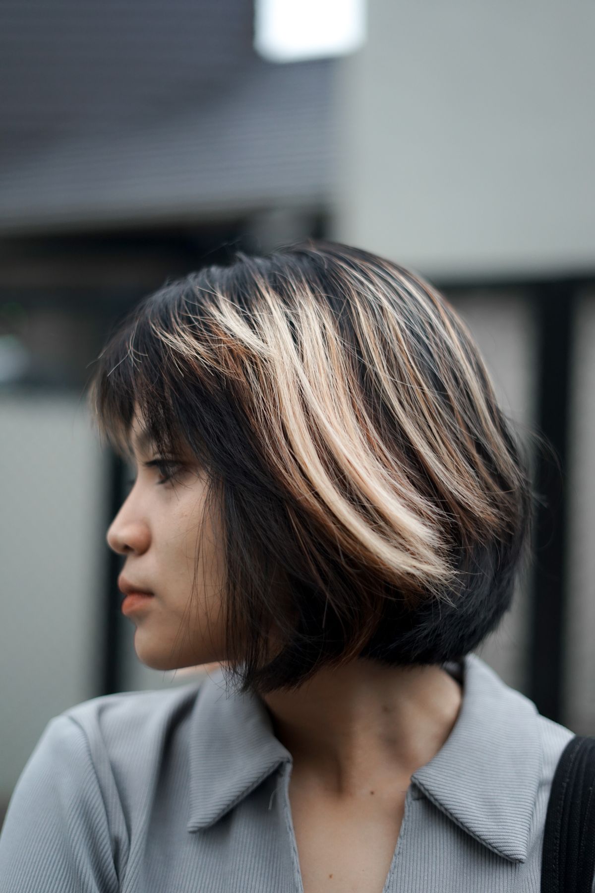 Woman in side profile with a dark bob, a short fringe and bright blonde face framing streaks, wearing a grey collared shirt