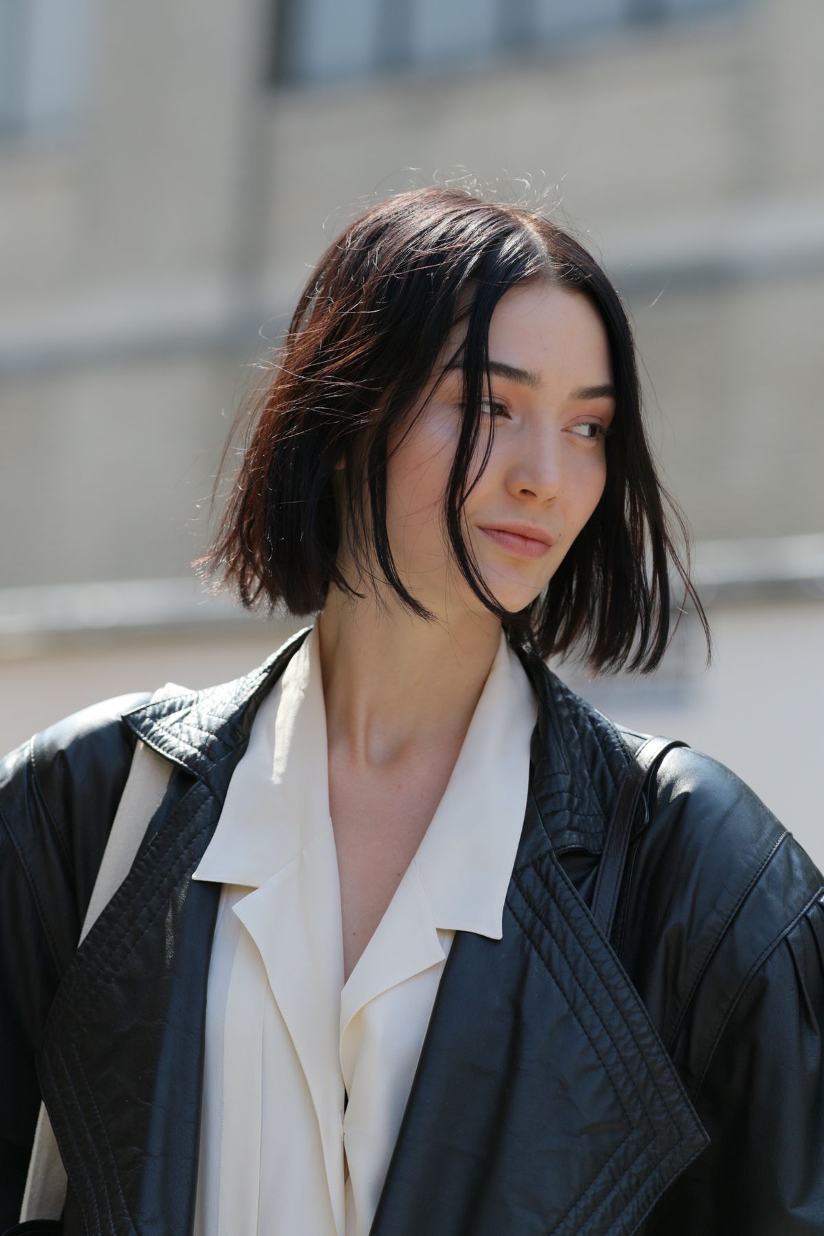 Woman with a jet black tousled bob and a centre part, wearing a black leather jacket over a white blouse in front of a building
