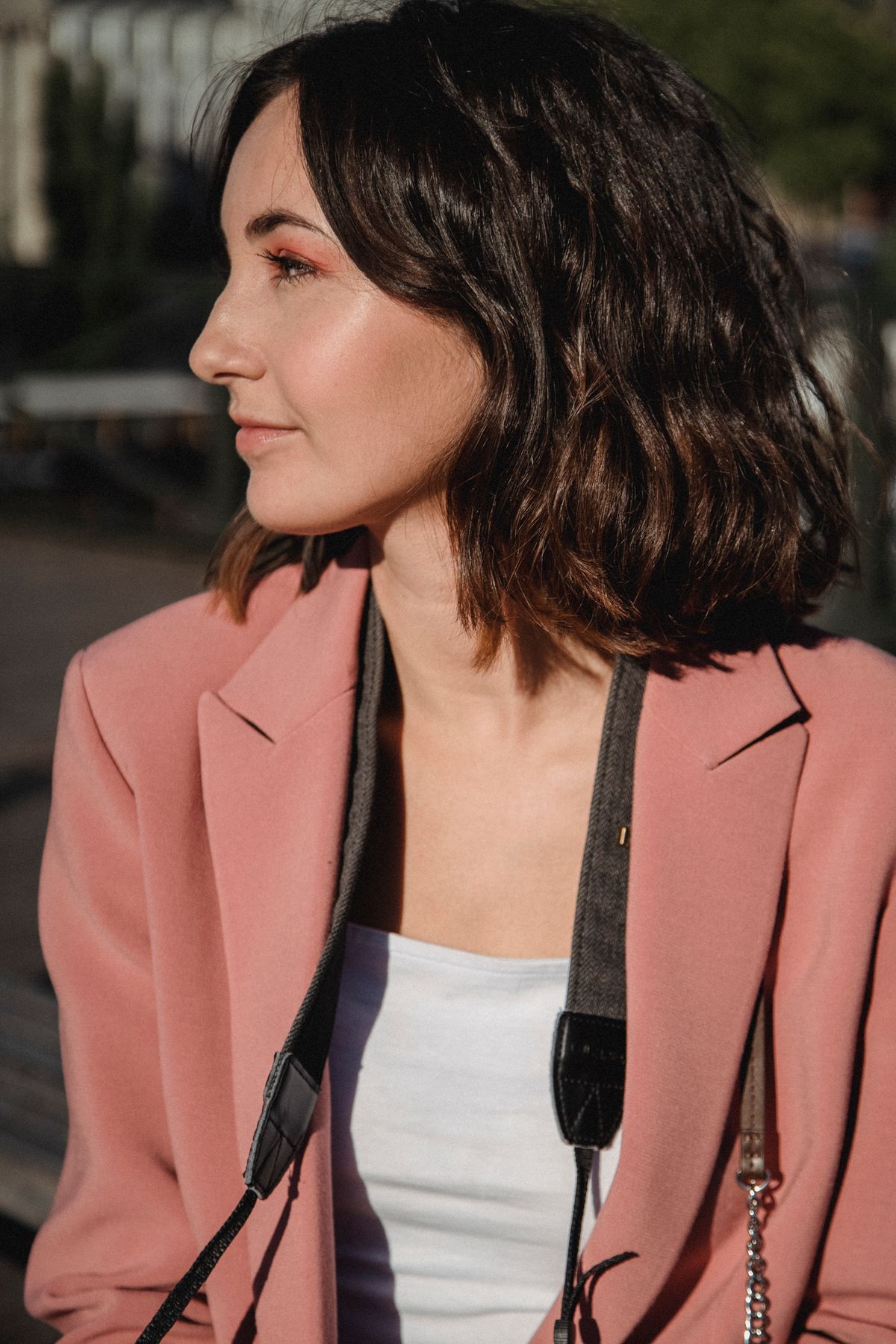 Woman in profile with a wavy dark brown bob grazing her shoulders, wearing a pink blazer with a camera strap over her shoulder