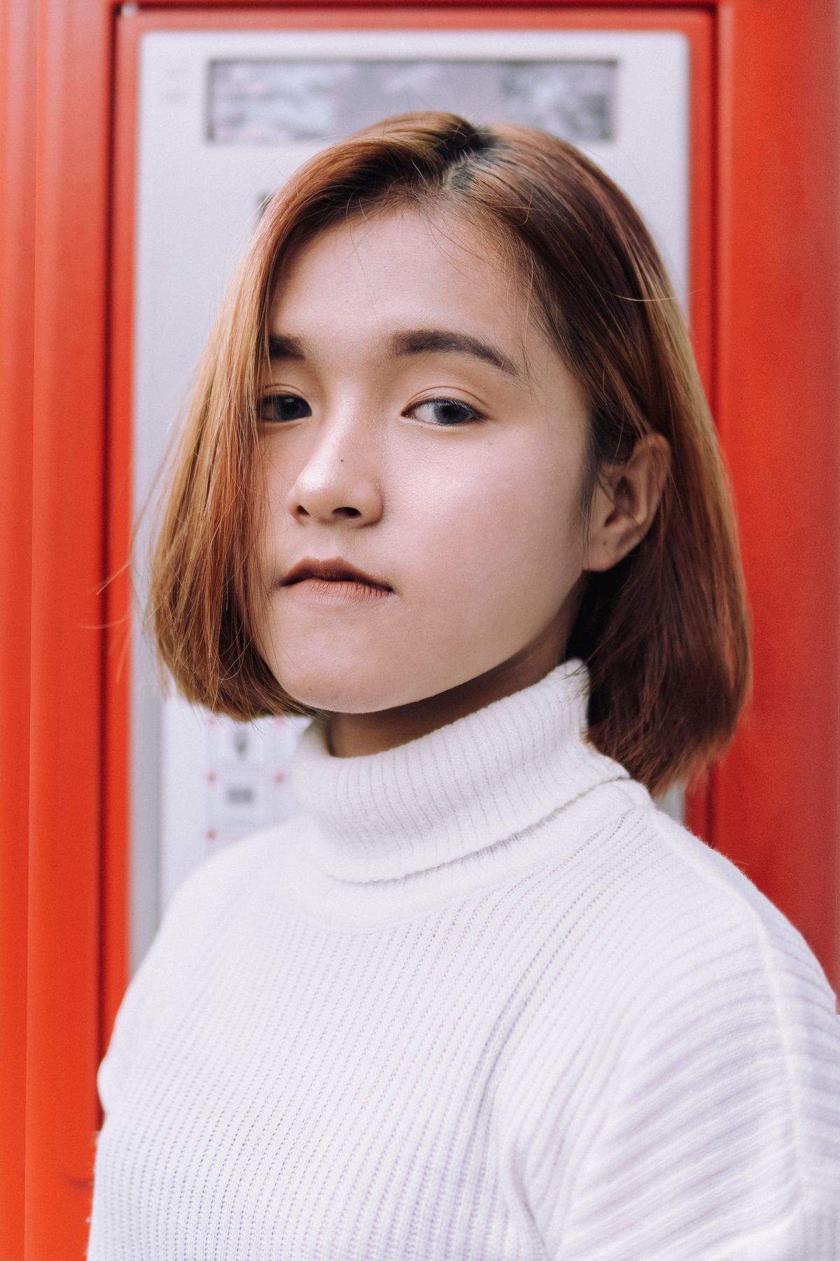 Woman with a sleek chin length copper auburn bob and a side part, wearing a white turtleneck in front of a red phone box