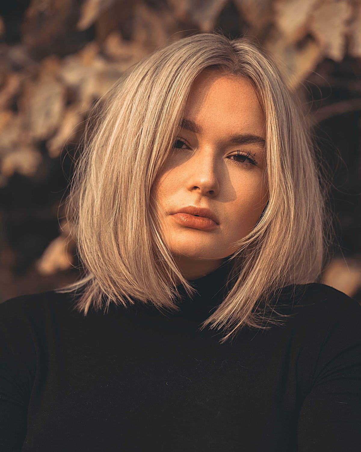 Woman with a sleek chin length blunt bob in warm honey blonde, wearing a black turtleneck against a golden autumn background
