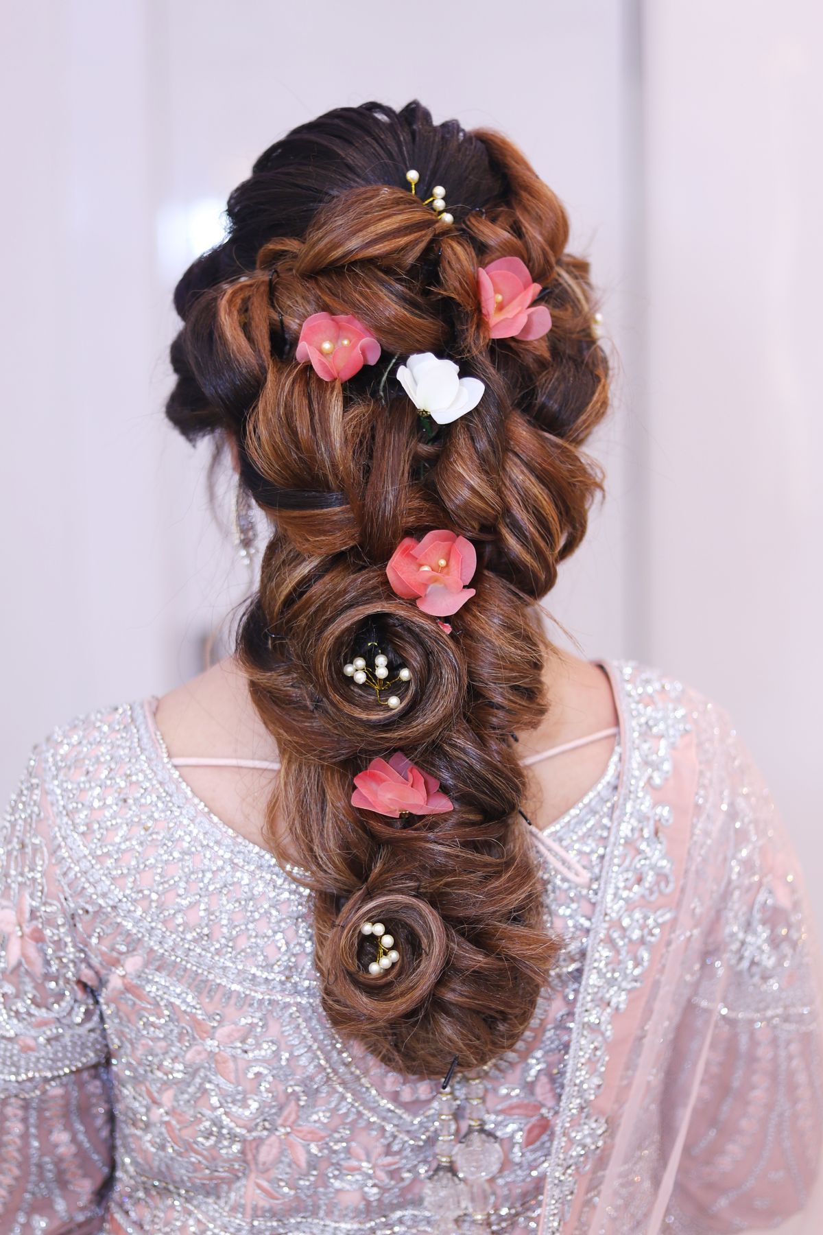 Back view of a woman's long chestnut hair in a loose, voluminous braid decorated with pink and white flowers and small pins, wearing a pale embroidered outfit