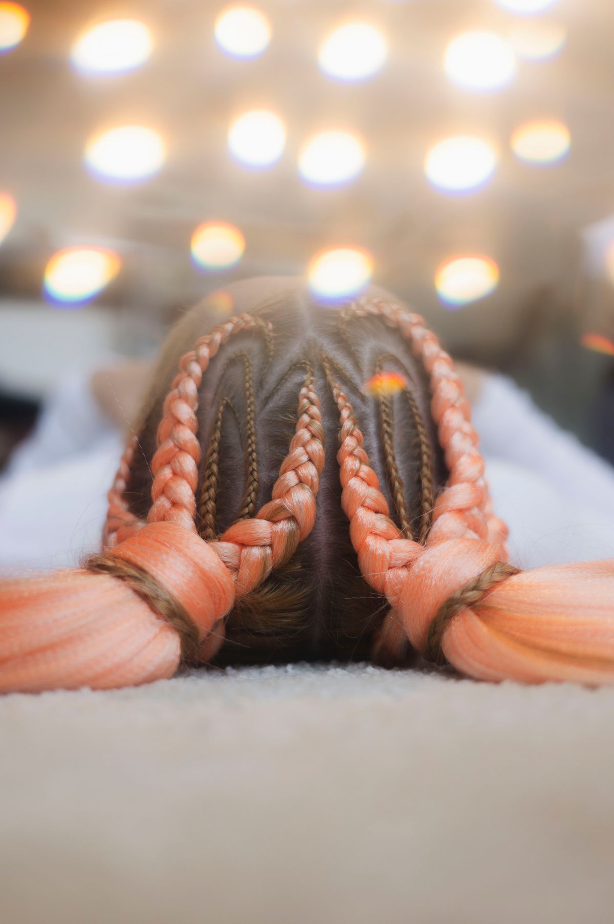Woman lying face down seen from above with dark cornrows leading into long braids with coral pink extensions, with soft lights in the background