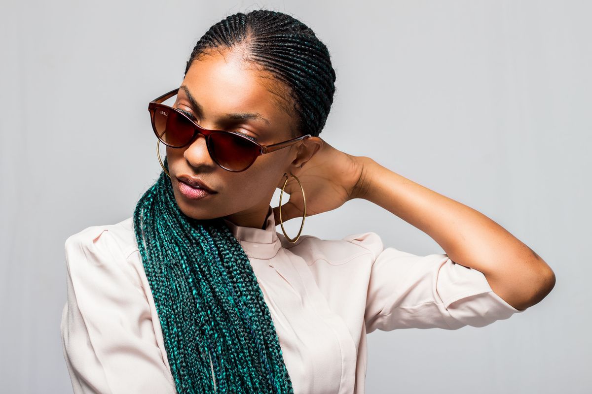 Woman in red tinted sunglasses and a hoop earring with fine cornrows pulled back and long teal green braids over one shoulder, wearing a cream blouse