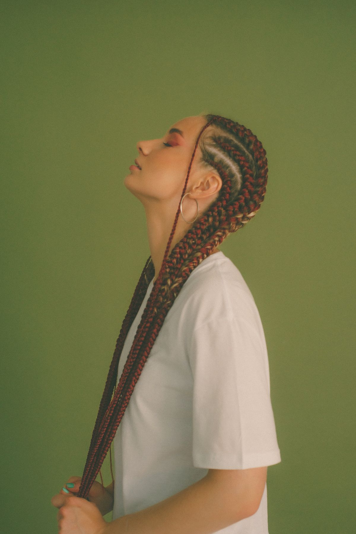 Woman in profile with her head tilted back, holding long cornrow braids with dark red extensions, wearing a white t-shirt against an olive green wall