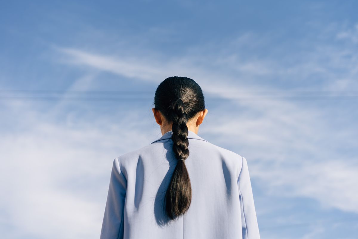 Woman seen from behind against a blue sky wearing a light blue blazer and her dark hair in one long, neat braid down her back
