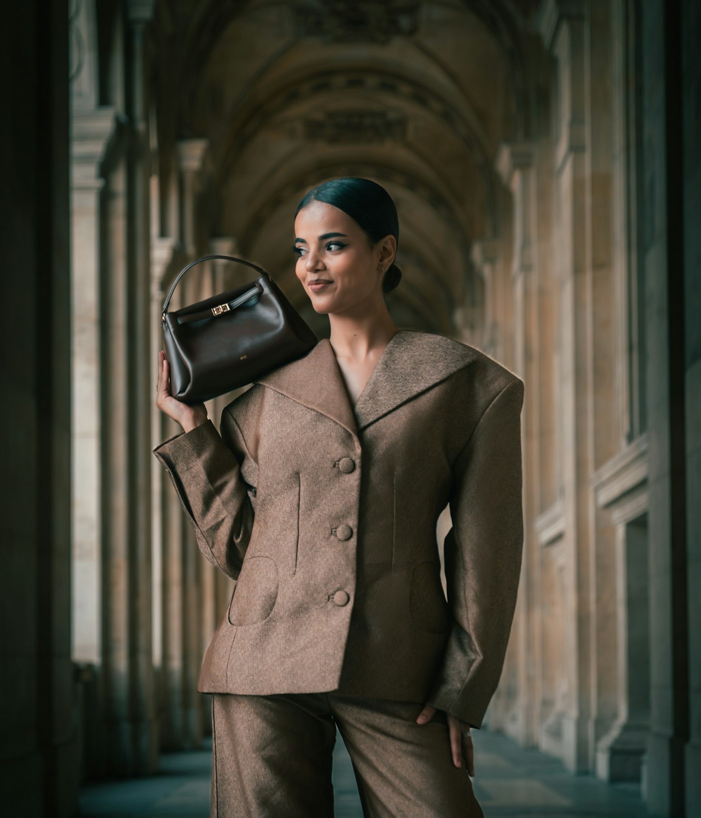 Woman in a brown suit holding a dark handbag in a stone corridor