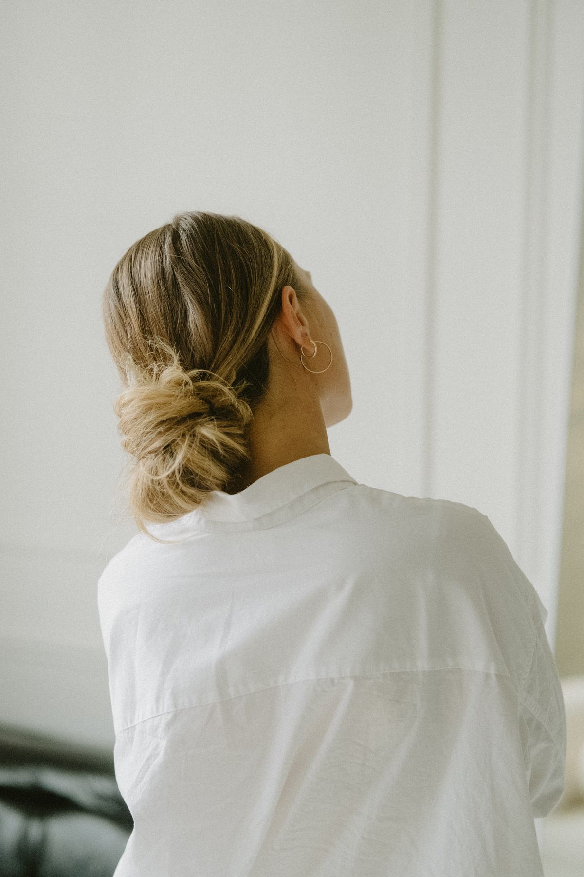 Woman with blonde hair seen from behind wearing her hair in a loose, textured low bun at the nape of her neck, in a white oversized shirt