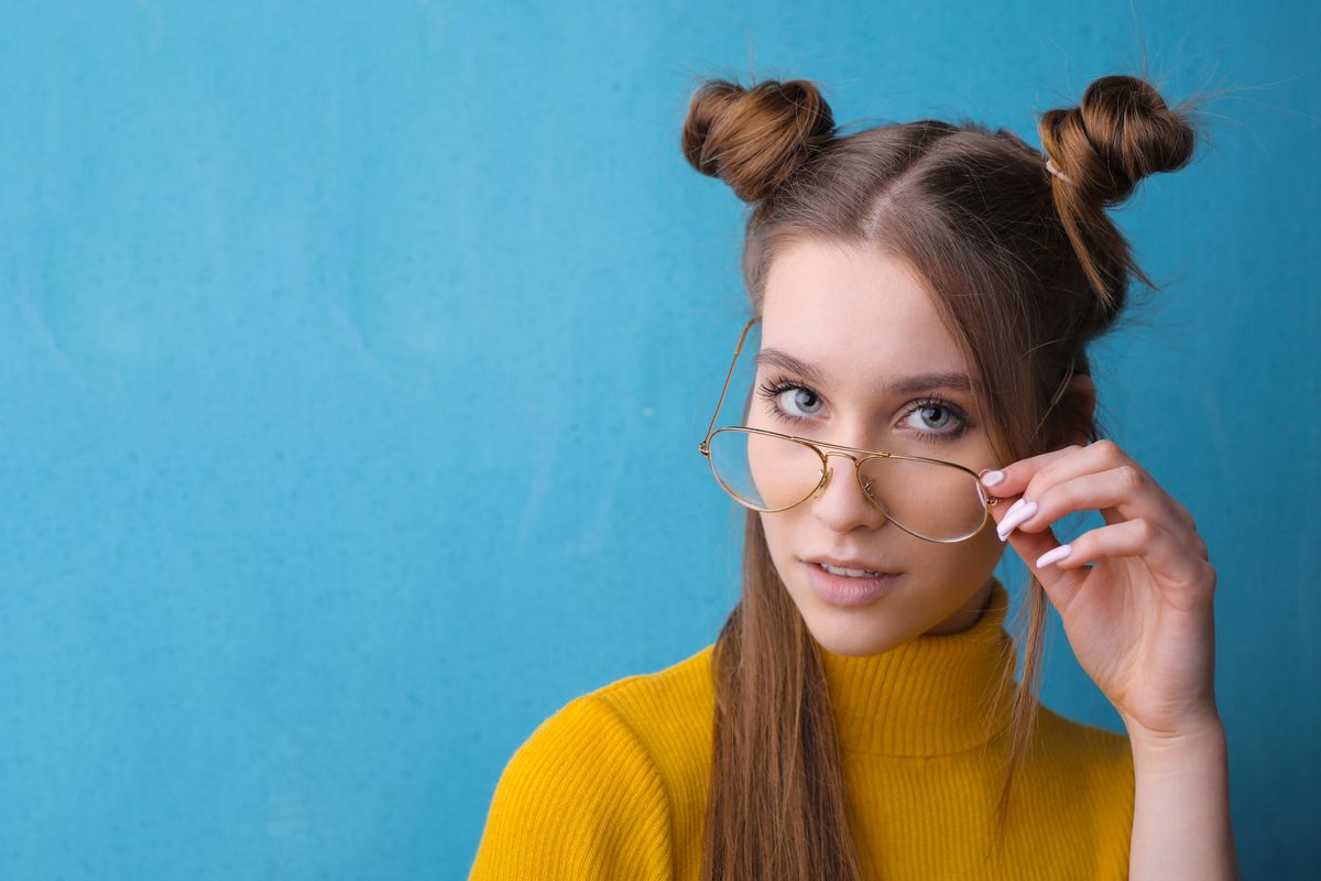 Woman wearing round glasses and a mustard yellow turtleneck with her long, straight hair in two small buns on top of her head against a blue wall