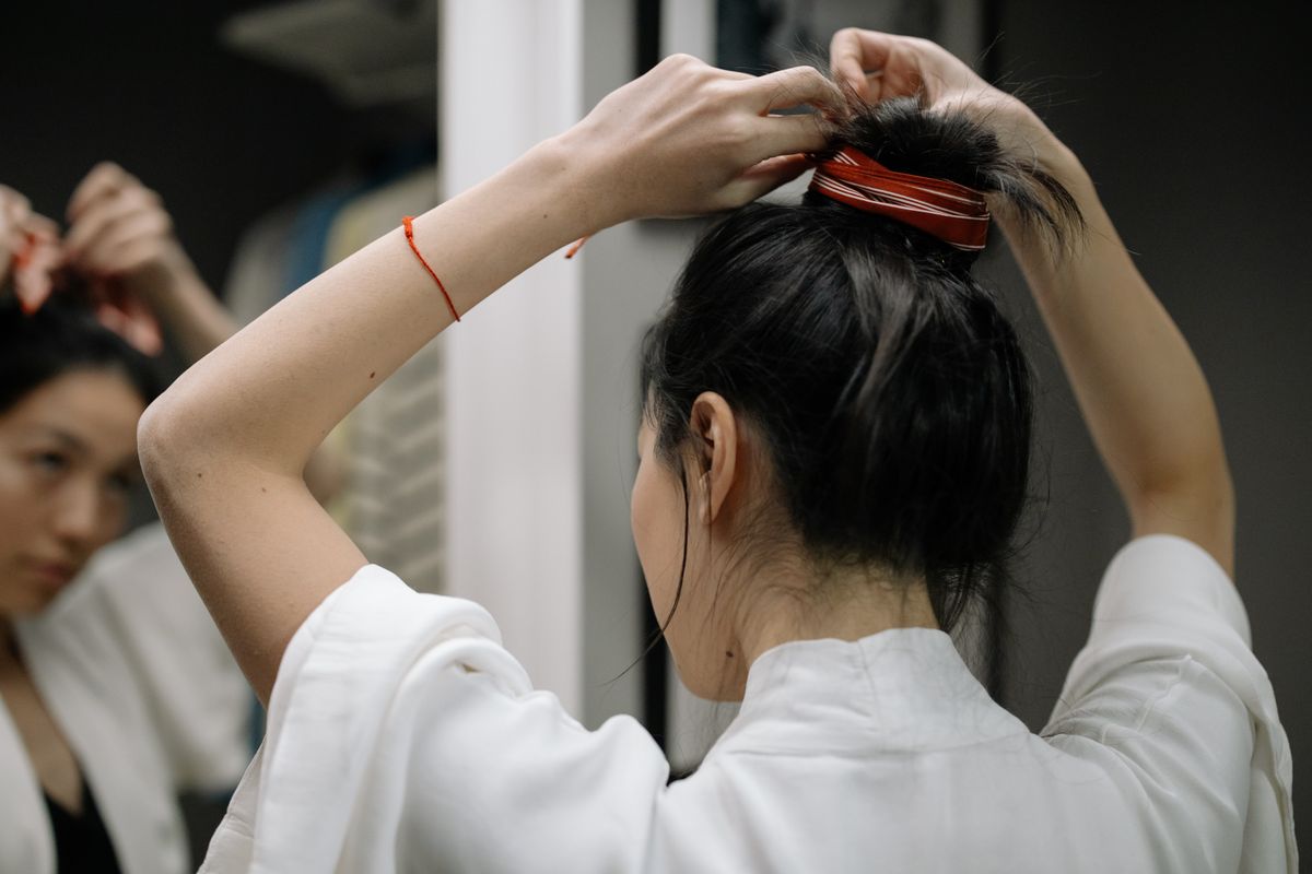 Woman with dark hair raising both arms to pull her hair into a high bun and secure it with a red hair tie, in front of a mirror and wearing a white top