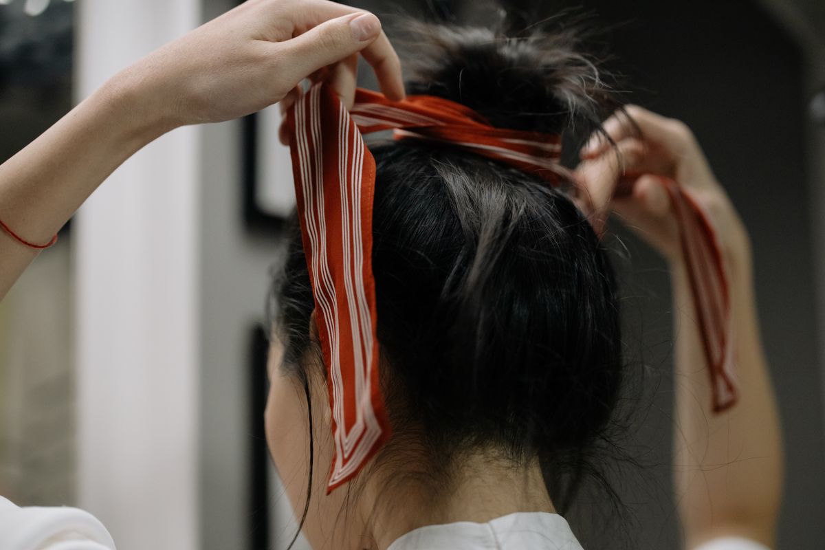 Woman tying a red and white striped scarf around her dark hair bun, hands raised, wearing a white top