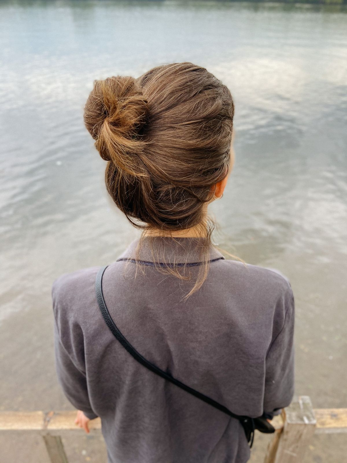 Woman seen from behind beside a lake, wearing her light brown hair in a messy high bun and a grey lilac top with a bag strap over her shoulder