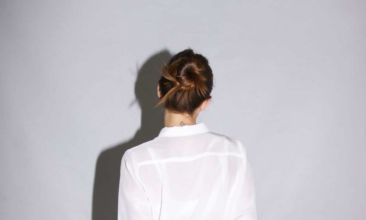 Woman seen from behind in a sheer white shirt against a pale grey wall, with her reddish brown hair twisted into a small, neat bun at the nape