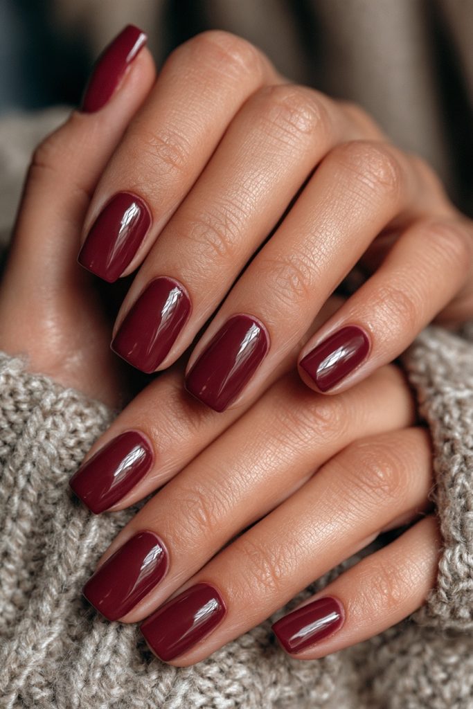 Hands in a grey knit sweater with short square nails in glossy burgundy polish