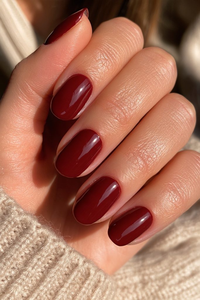Hand resting on a cream knit sweater with short oval nails in glossy dark burgundy polish
