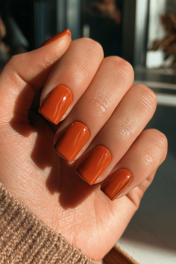Hand in sunlight showing short glossy burnt orange square nails and a taupe knit sleeve