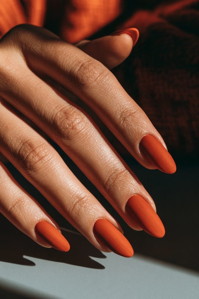 Long matte burnt orange almond nails on a hand resting against a dark knit sleeve