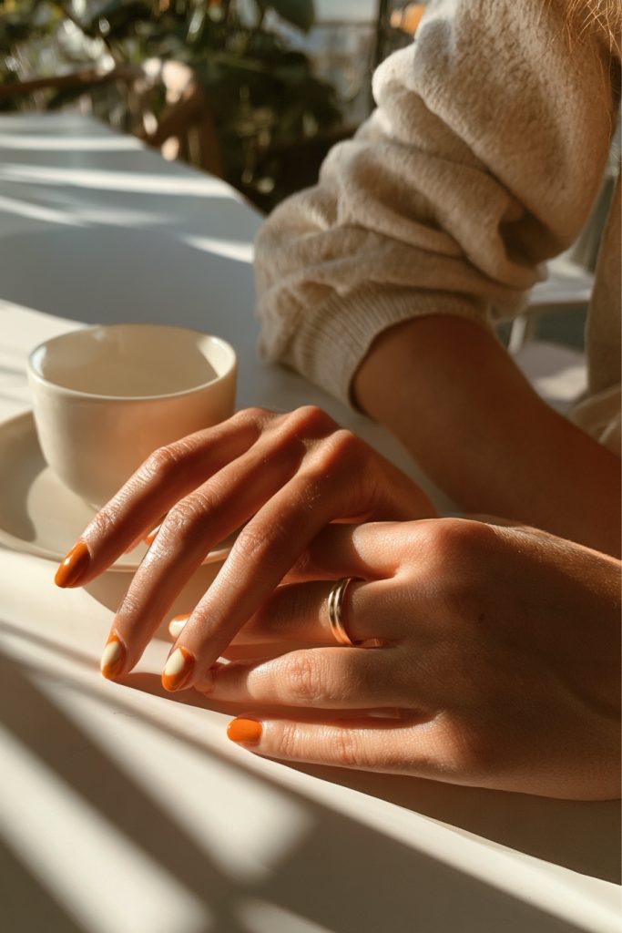 Hands resting on a white table beside a white cup, with short rounded burnt orange nails and a cream sweater sleeve