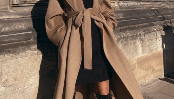 Woman in a belted camel wool coat over a black turtleneck dress with black knee high boots in front of a stone building