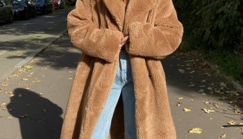 Woman in a long camel faux fur teddy coat with light blue jeans on a leafy street