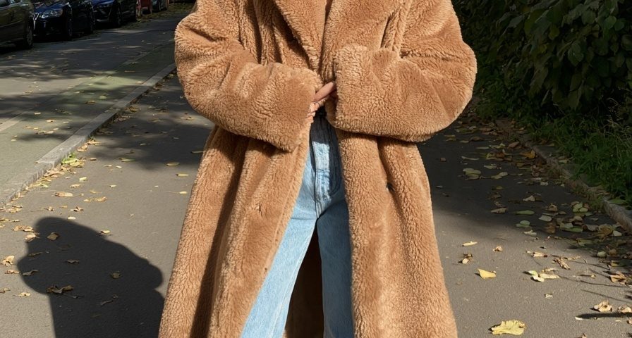 Woman in a long camel faux fur teddy coat with light blue jeans on a leafy street