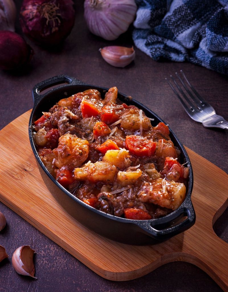 Dark cast iron pot of slow-cooked stew with tomatoes, potatoes and vegetables on a wooden board