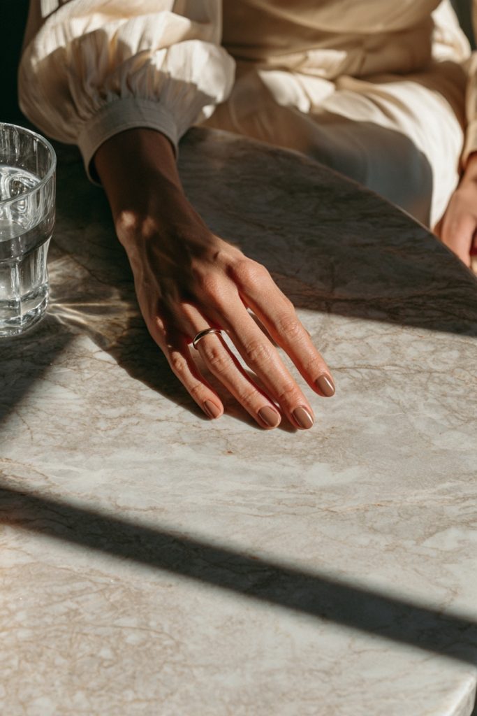Hand resting on a marble table beside a glass of water with dark chocolate brown nails and a white puffed sleeve