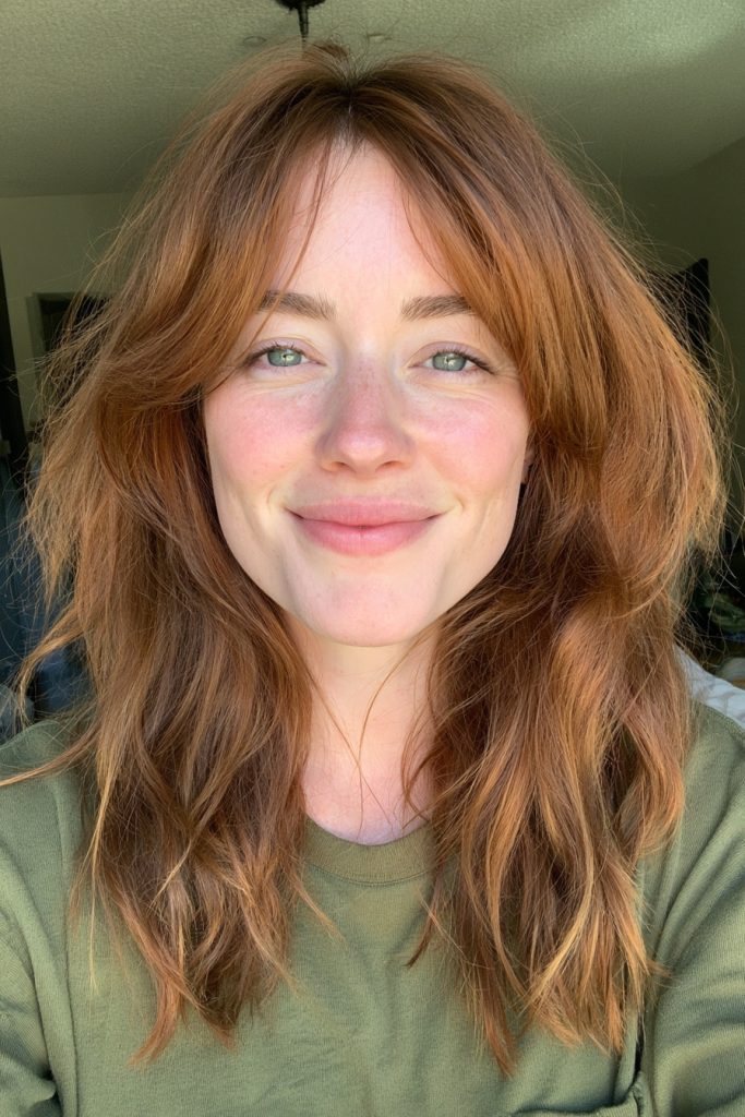 Close up of a woman with medium length copper hair, curtain bangs and freckles wearing an olive green top