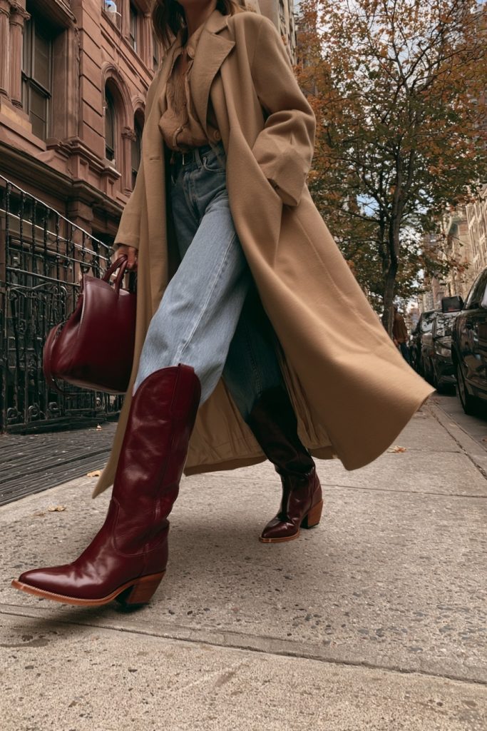 Close up of burgundy leather cowboy boots with light wash wide leg jeans, a long camel coat and a burgundy leather bag