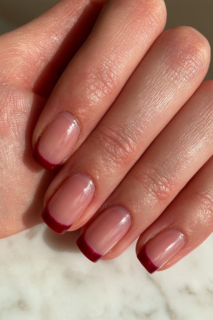 Close-up of a hand with short sheer pink nails and thin cranberry red French tips on a marble surface